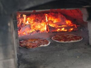 two pizzas cooking in a woodfired pizza oven. glowing coals with a bit of fire in the back. Two pizzas with the cheese caramelized and the dough puffing up in the front.